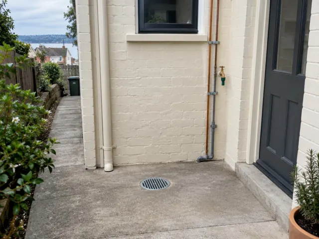 Outdoor courtyard gully with grate set in concrete beside a house wall and door in Neutral Bay