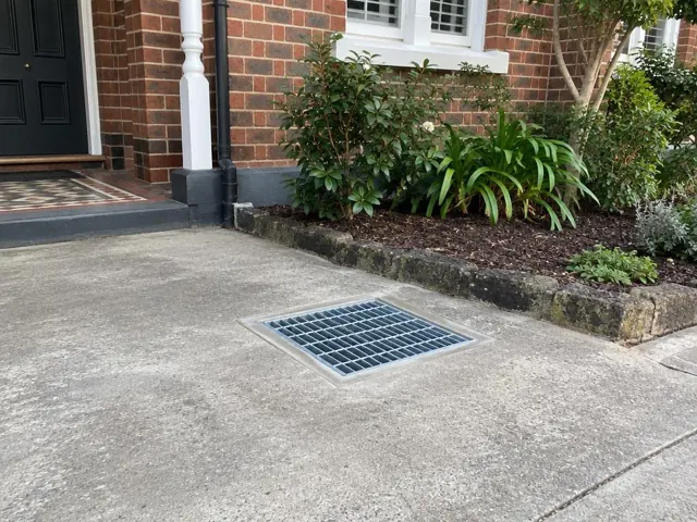 Cleared stormwater grate set flush in a concrete driveway beside a brick house in Neutral Bay